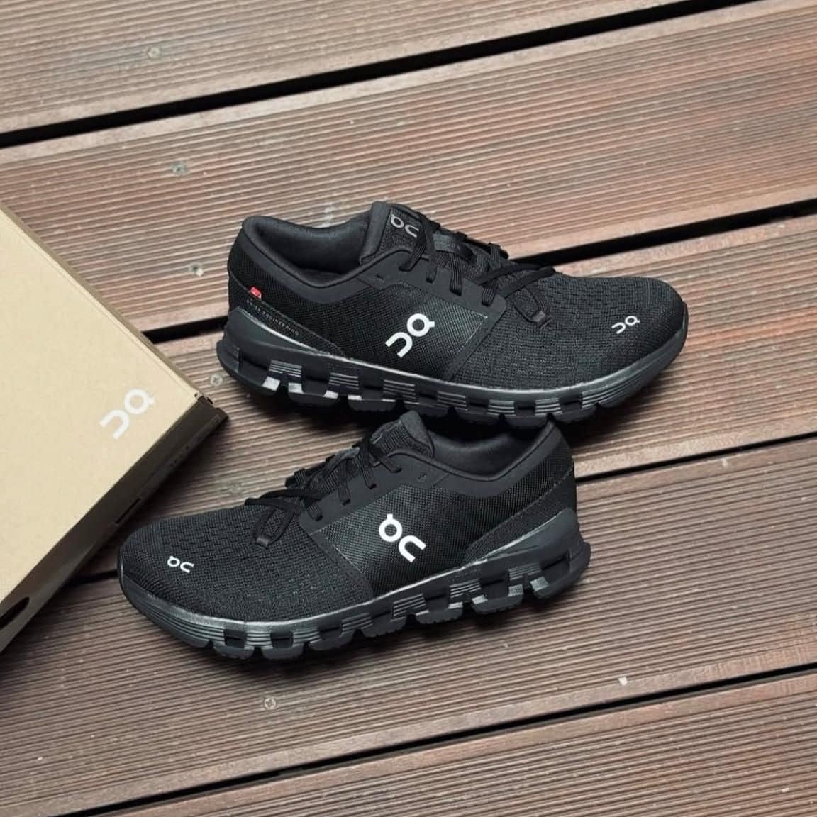 9b6df0be-8b0c-4b3f-80cf-f05a521c5e17 | AE JERSEY Pair of black On running shoes displayed on a wooden deck beside a shoe box.