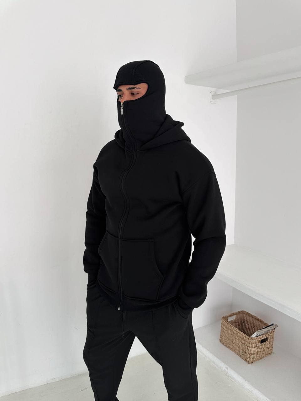 9c6b1411-3197-4c48-8dab-ddaf4edc2aa9 | AE JERSEY Person wearing a black balaclava and zip-up hoodie with hands in pockets, standing in a white room.