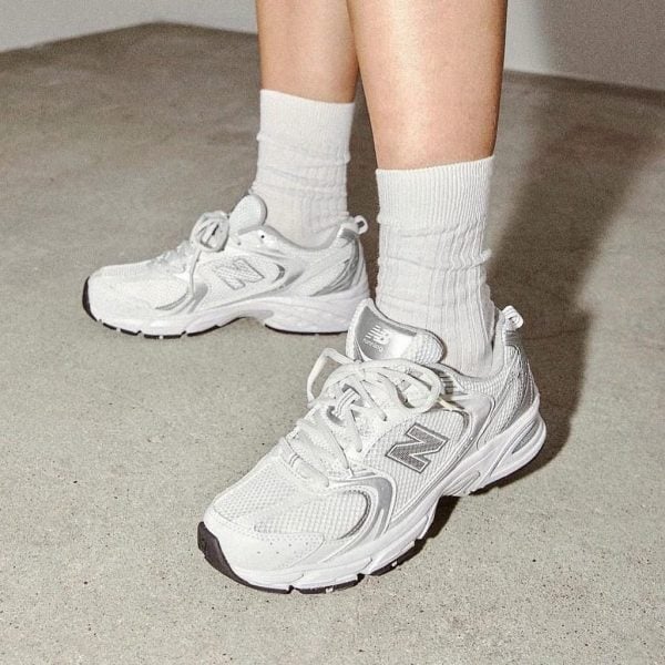Close-up of white New Balance running shoes on a person’s feet, wearing white crew socks, standing on a concrete floor.