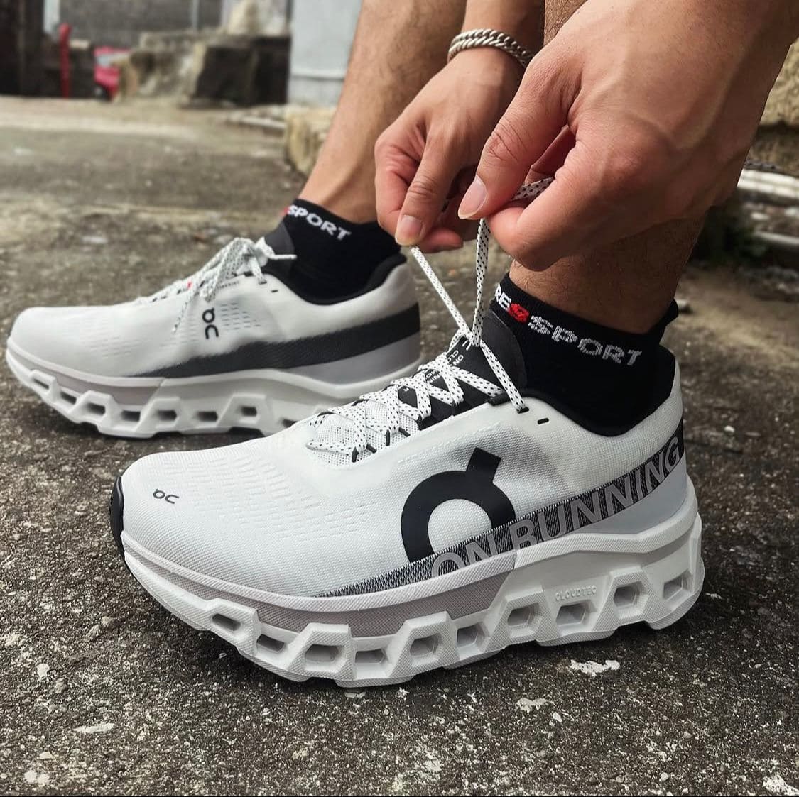 9dac2c3a-85a7-4a70-a041-bbe51ae88844 | AE JERSEY Person tying white On Running sneakers with chunky soles, black SPORT socks, outdoors on concrete surface.