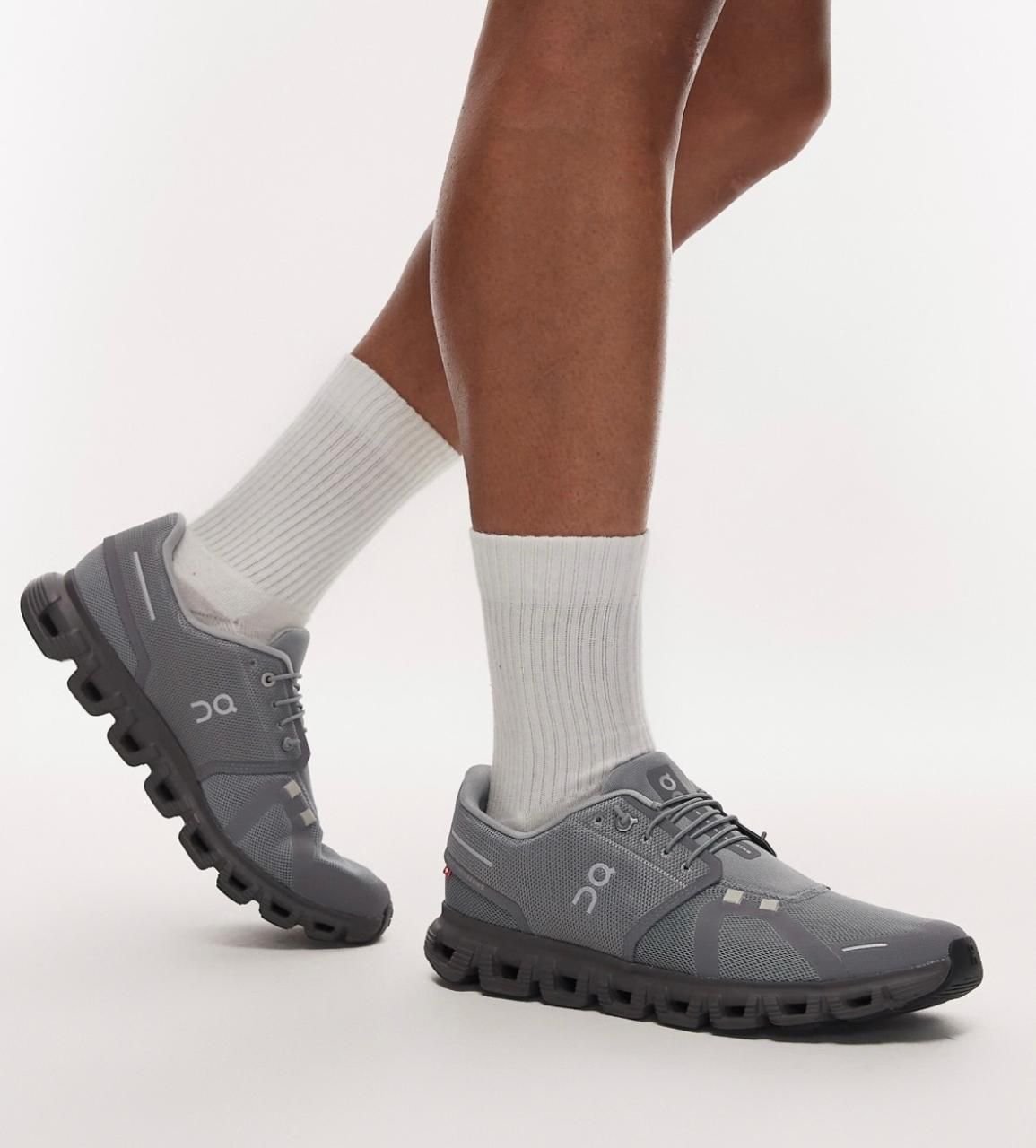 9e1fc904-2e56-4b25-81d3-7659f0880974 | AE JERSEY Gray athletic shoes with a chunky segmented sole, worn with white crew socks during a mid-step pose.