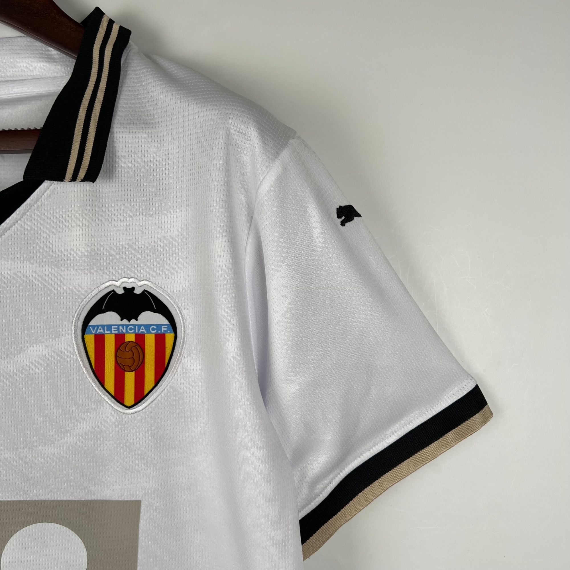 9f9fa105_532ca83c-ce47-4244-b3e7-8063d3e51608 | AE JERSEY Close-up of a white Valencia CF soccer jersey with the club crest on the chest and black-gold striped collar detail.