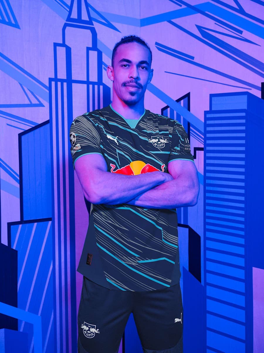 AKrF6JG10NJ6avY | AE JERSEY Athletic man with arms crossed stands in front of a blue neon city mural, wearing a dark jersey with teal stripes and a Red Bull logo.