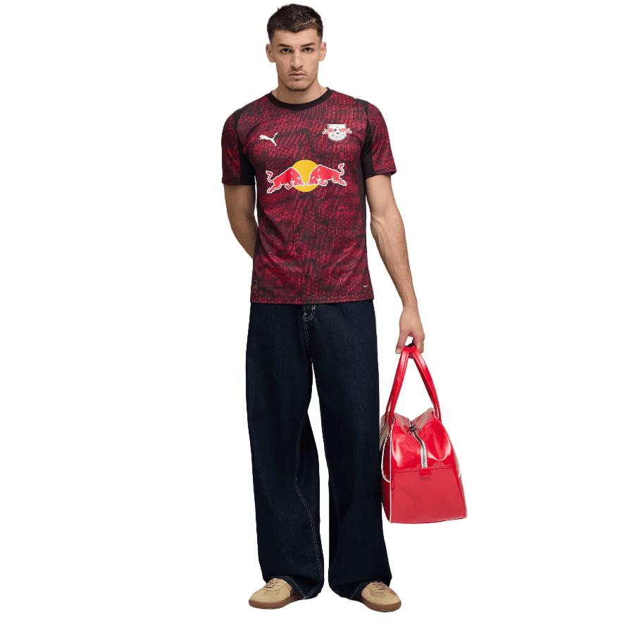 AQ | AE JERSEY Male model wearing a maroon RB Leipzig-style soccer jersey with Red Bull logo, dark jeans, tan shoes, and holding a red handbag.