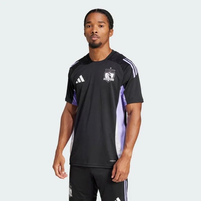 Camiseta_Entrenamiento_Colo-Colo_672x672_bf385a35-fd28-48cb-915a-bad9909dc260 | AE JERSEY Man modeling a black Adidas soccer jersey with white stripes on the shoulders and purple side panels against a light background.