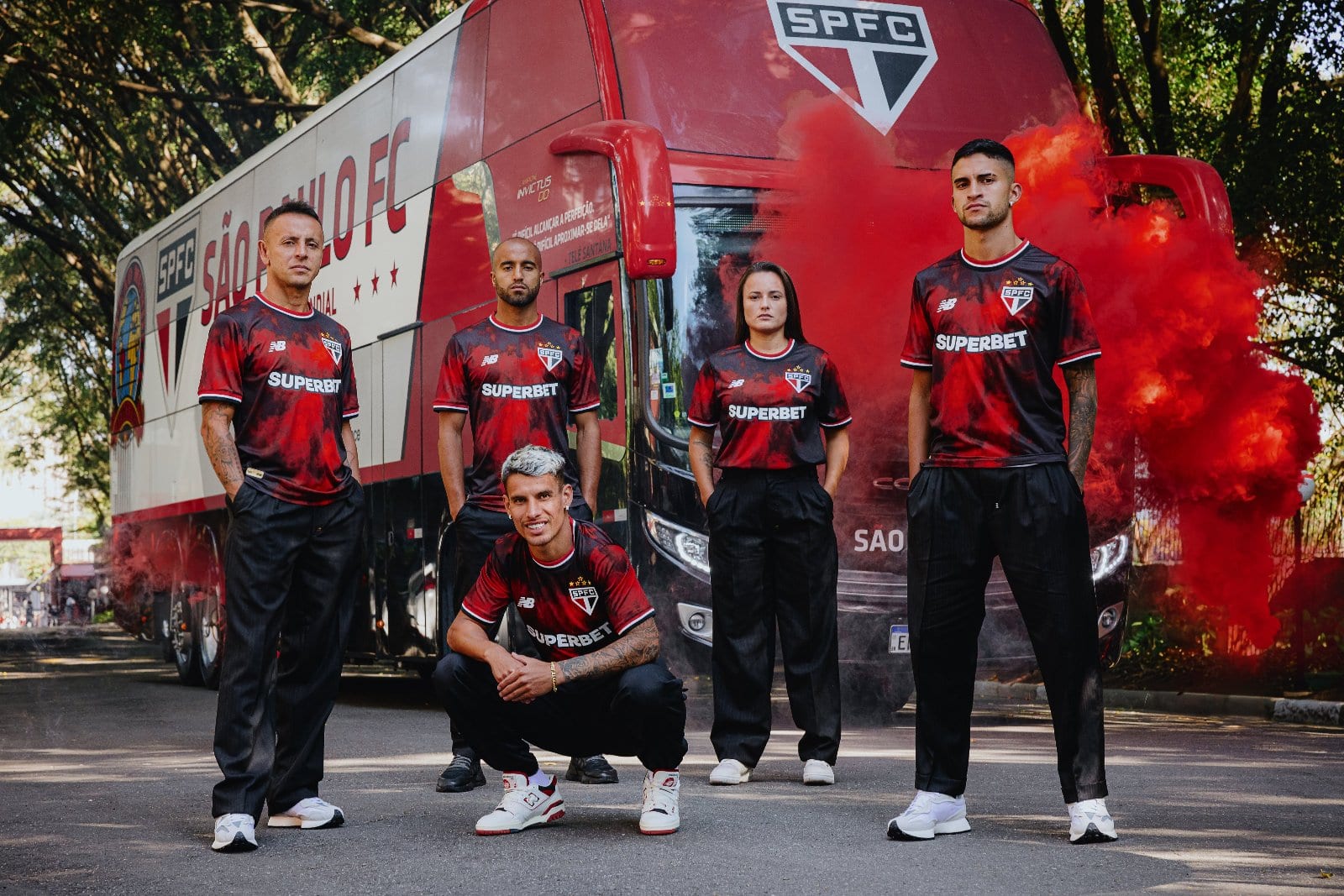 FIZM4gWSvJO2R2k | AE JERSEY Five soccer players in red-and-black SPFC jerseys pose in front of a team bus, red smoke billowing nearby, outdoors.