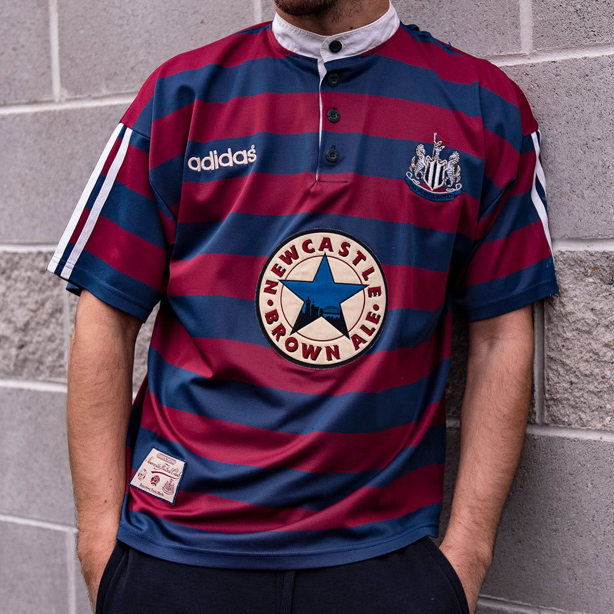FlI_ZFLX0AAuKRI | AE JERSEY Man wearing a Newcastle United jersey with maroon and blue horizontal stripes, Adidas logo, team crest, and a large Newcastle Brown Ale badge on the chest.