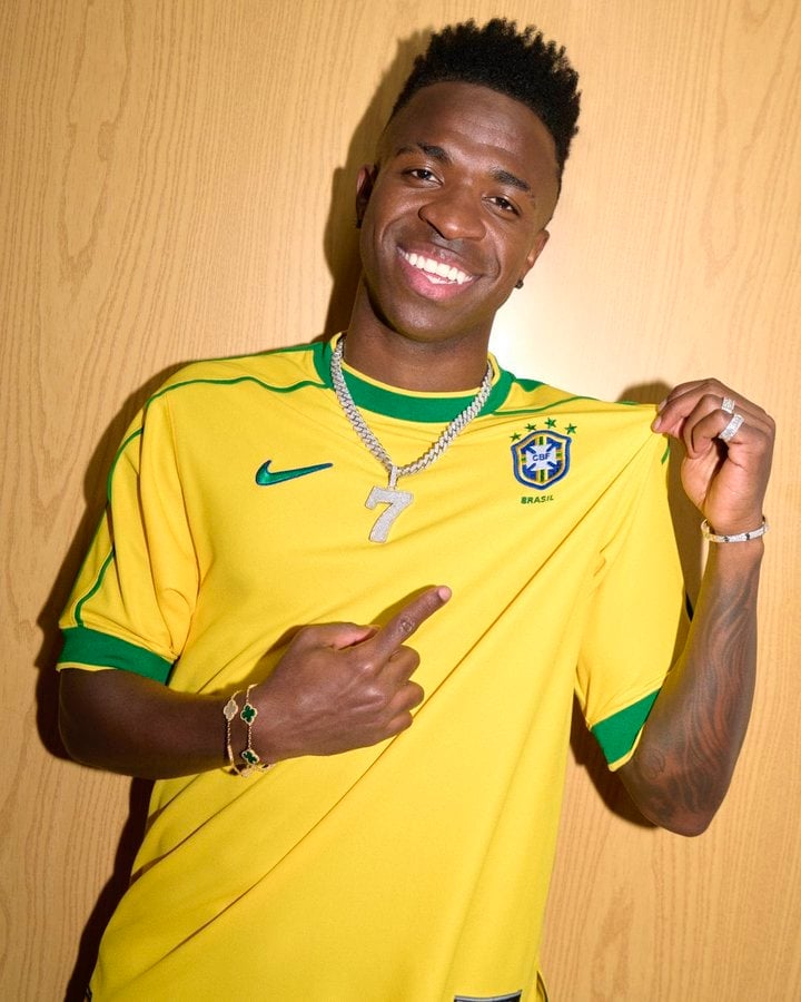 GQmVUXEWIAAmuik | AE JERSEY Smiling man in a Brazil soccer jersey points to a silver '7' pendant on his chain against a wooden background.