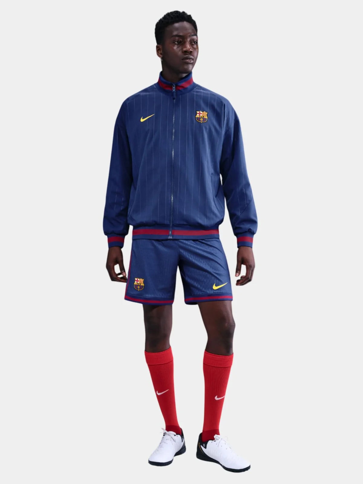 GT | AE JERSEY Man wearing FC Barcelona blue training jacket and shorts with red accents and white cleats, standing against a light background.