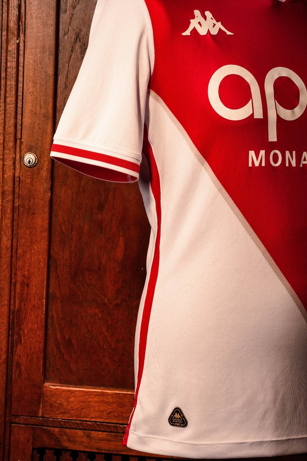 GvHAbflyHaShHSz | AE JERSEY Close-up of a red and white sports jersey hanging against a wooden door, featuring the Kappa logo on the shoulder and partial text/logo on the front.
