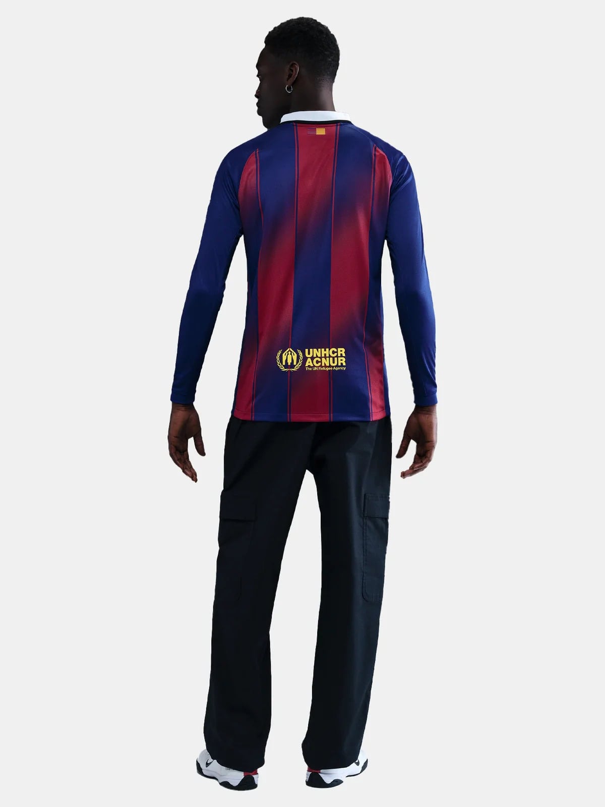 HJ4611-456-PHSBMG01_copia | AE JERSEY Back view of a man in a blue and red striped long-sleeve shirt with UNHCR logo on the lower back, wearing black cargo pants and white sneakers.