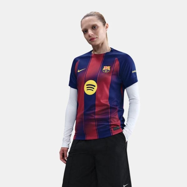 Young woman wearing FC Barcelona home jersey with blue and red vertical stripes, yellow Spotify logo, and Nike swoosh, standing with hands in pockets against a light backdrop.