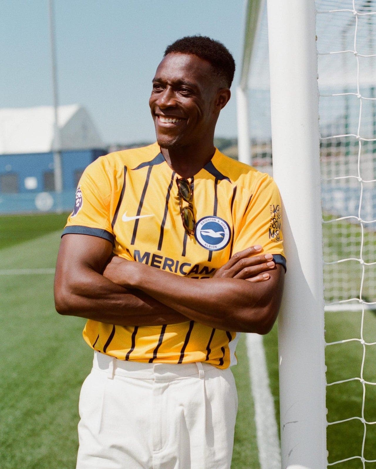 J0ovUj5yfskvsk8_1 | AE JERSEY Smiling man in a yellow Brighton & Hove Albion soccer jersey leaning against a goalpost on a sunny outdoor field.