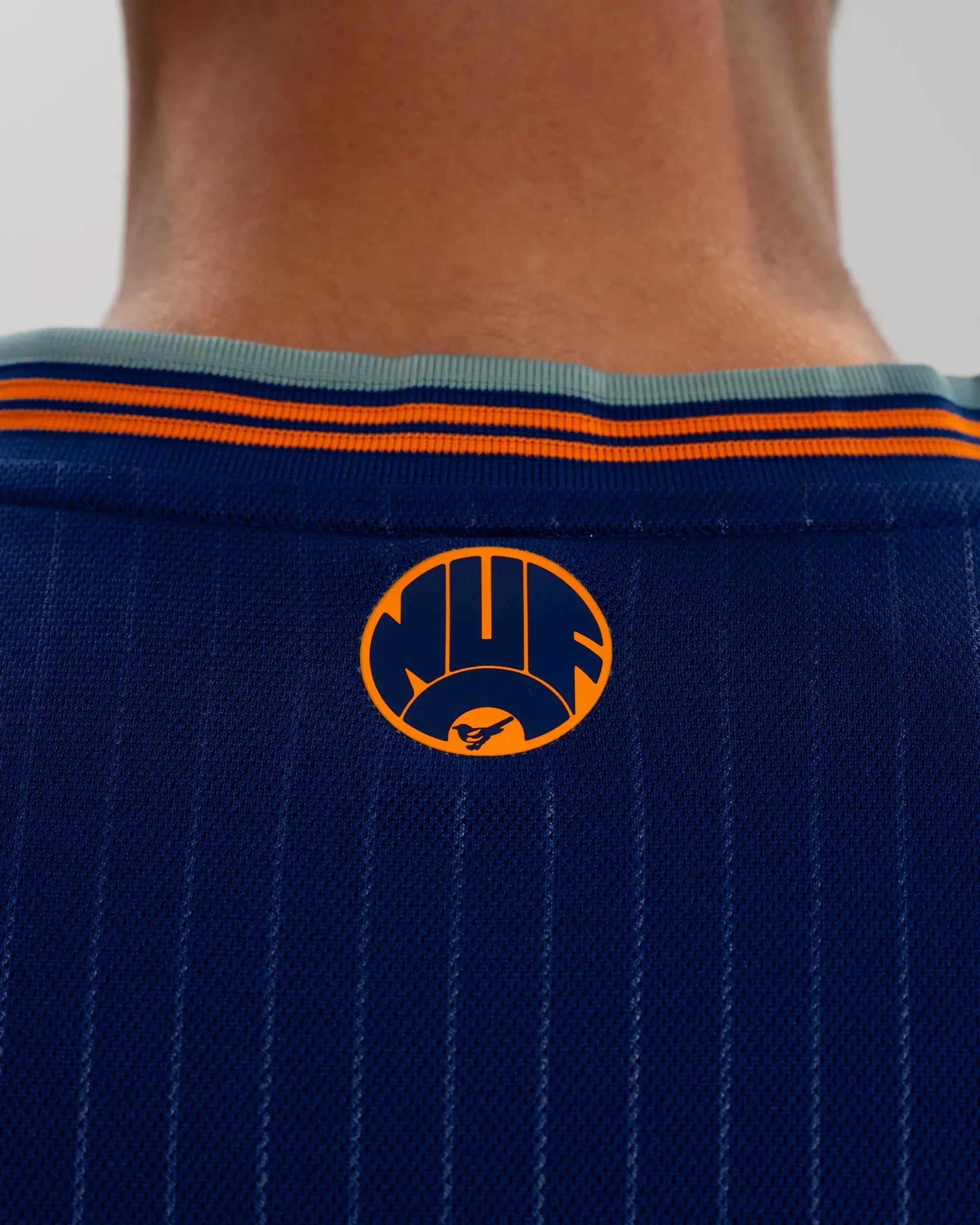 JI7388_MOD_A_ECOM_13 | AE JERSEY Back view of a person wearing a dark blue athletic jersey with orange stripes and a circular orange logo on the back collar area, neck visible.