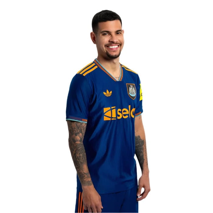 JI7388_PLAY_BG_11 | AE JERSEY Man in a blue soccer jersey with orange accents, smiling and standing sideways, tattoos visible on both arms.