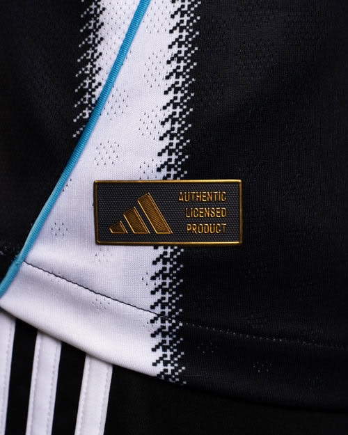JI7391_MOD_26_77f8f18d-03f8-4e3a-b093-6408b0c509b8 | AE JERSEY Close-up of a rectangular black and gold badge reading 'AUTHENTIC LICENSED PRODUCT' pinned to a black and white athletic garment with a blue zipper edge visible in the background.