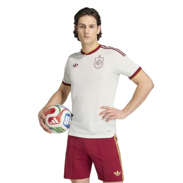 Male soccer player wearing a white Spain national team jersey with red trim, red shorts, and holding a colorful soccer ball against a white background.