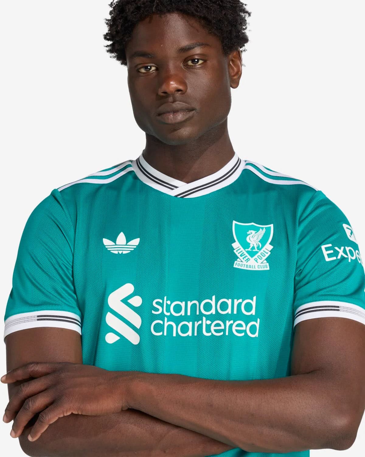 JV6427_7_APPAREL_OnModel_DetailView1_custom_1-lowres | AE JERSEY Young man with arms crossed wearing a teal Liverpool FC jersey featuring Standard Chartered and Adidas logos.