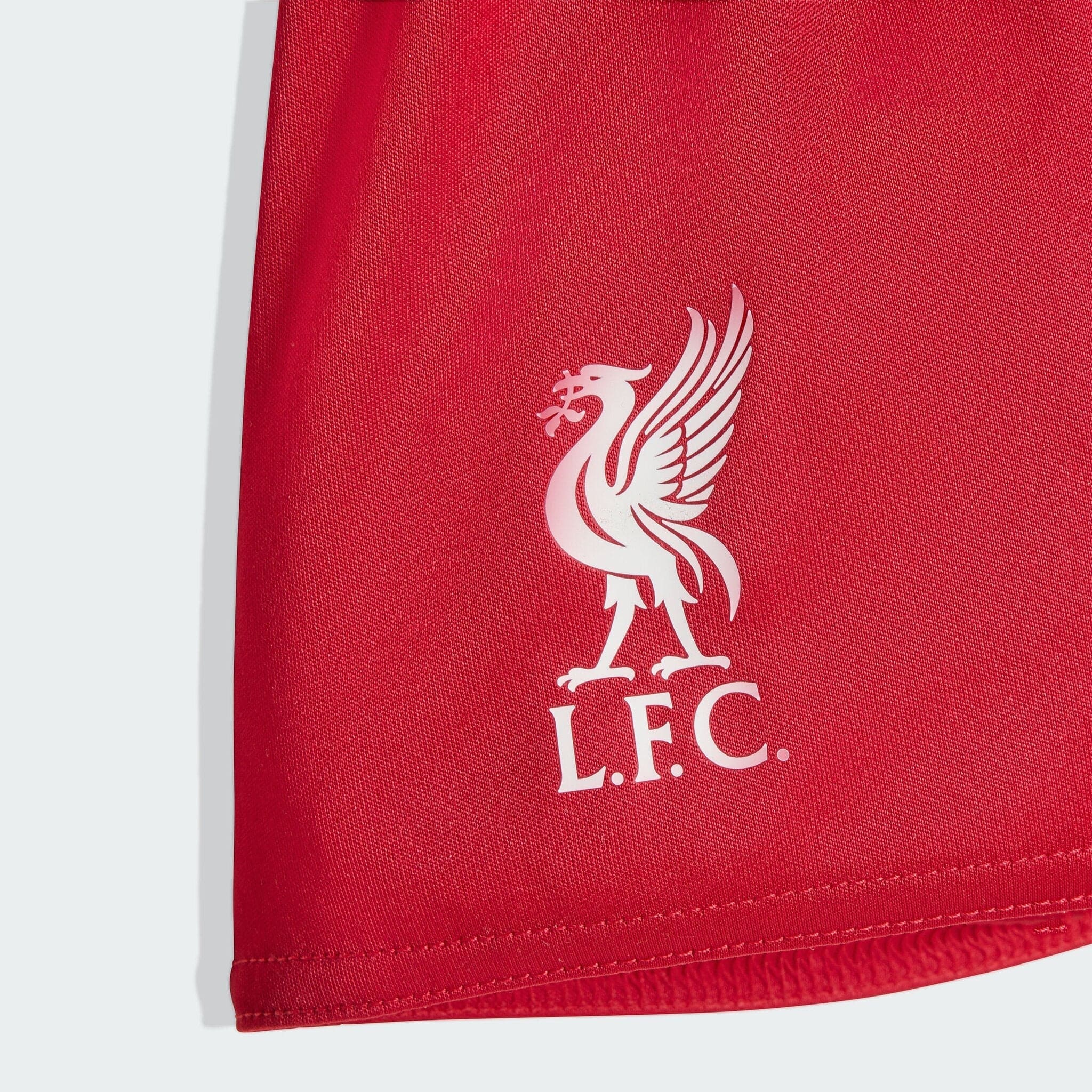 JV6443_D3_Flat_eCom-lowres | AE JERSEY Close-up of a red fabric surface featuring a white Liverpool FC crest partially visible.