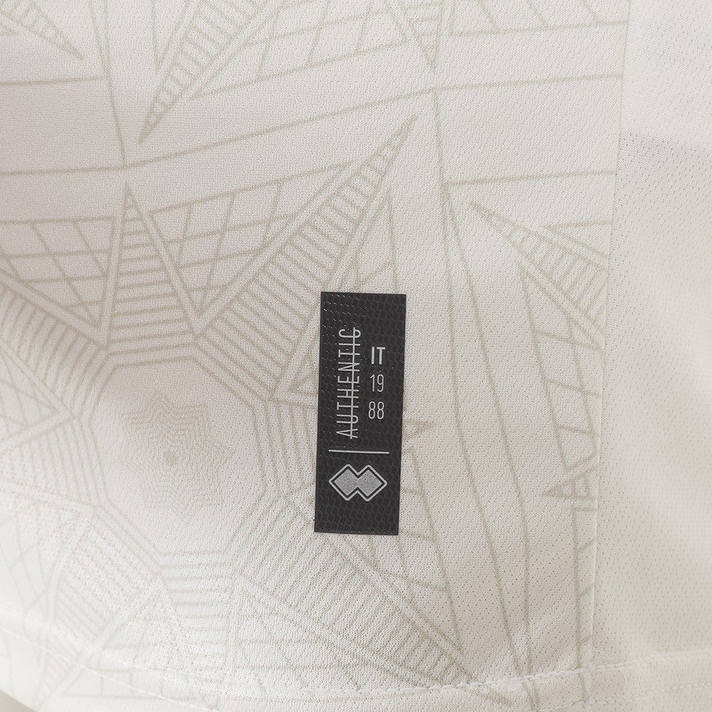 JlLqIAXVSn2nkhg | AE JERSEY Close-up of light patterned fabric with a small black rectangular brand tag featuring a white geometric logo.