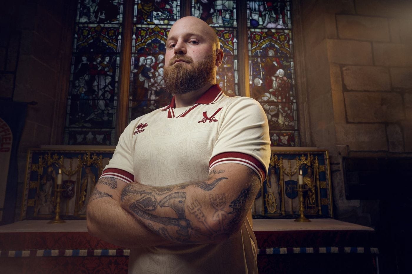 LnX4wu2sNXj6Q2h | AE JERSEY Bearded man in a white sports jersey with arms crossed, standing in a church with a stained-glass window behind him.
