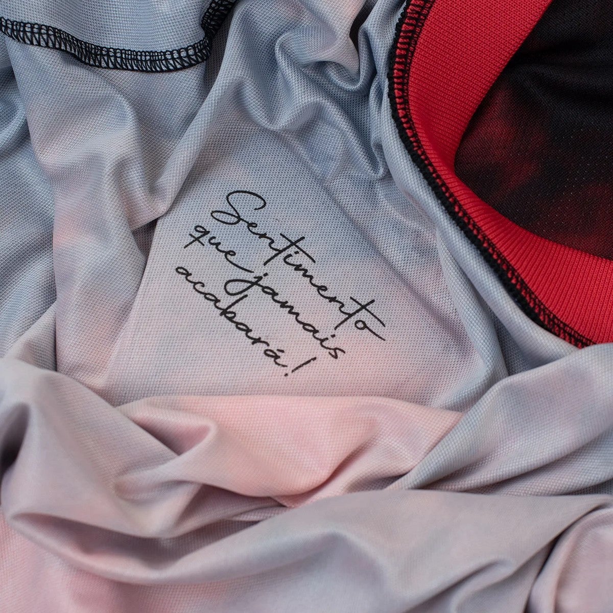 NrKt51pRKDF6LA0 | AE JERSEY Close-up of soft light-gray fabric with a handwritten cursive message, with pink fabric and a red-trim garment nearby.