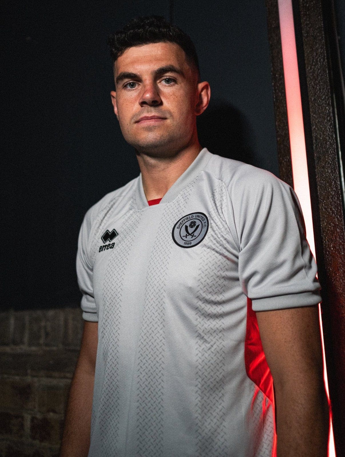 OY8PtsuB91l1KAE_0d73a6c7-cec2-4718-9d73-06b81baed519 | AE JERSEY Portrait of a man in a white football jersey leaning against a wall at night, red light illuminating his side.