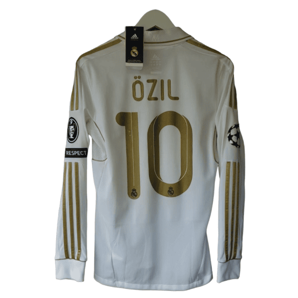 White Real Madrid football jersey with gold 'Özil' and number 10 on back, Champions League sleeve patch visible.
