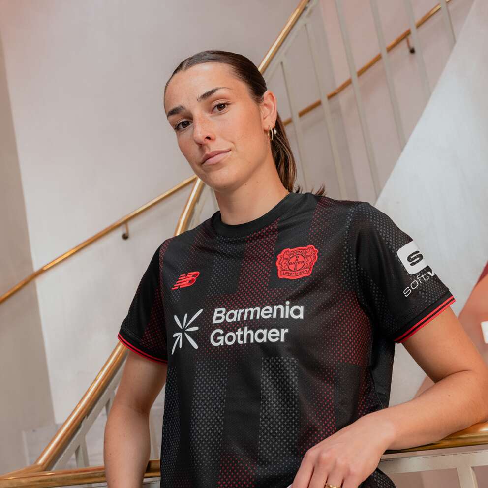 Portrait of a female football player in a black/red team jersey, leaning on a stair railing indoors, looking at the camera.