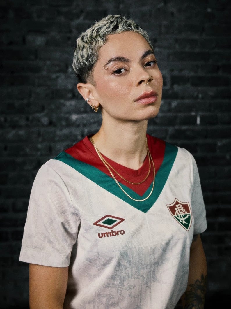 QgWMUa6sxl0gDrA | AE JERSEY Portrait of a person with short blond hair wearing a white soccer jersey with green and burgundy trim and crest, against a dark brick wall.