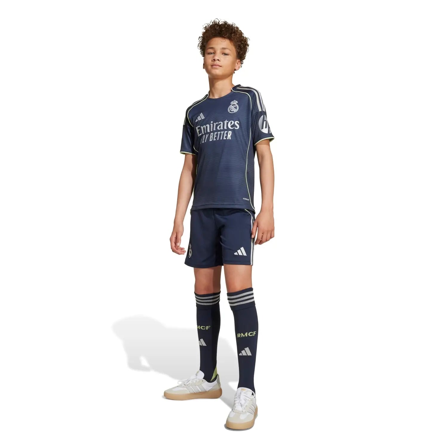 RMCFYO0095_8 | AE JERSEY Young boy standing in a navy Real Madrid kids football kit, jersey and shorts, with matching socks and sneakers, sponsor logo visible.