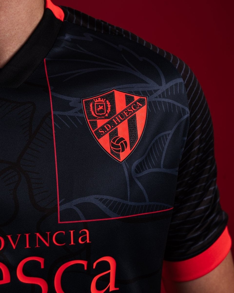 Rx8DGZnlFnAoNIv | AE JERSEY Close-up of a dark navy SD Huesca soccer jersey with a red crest and red trim.