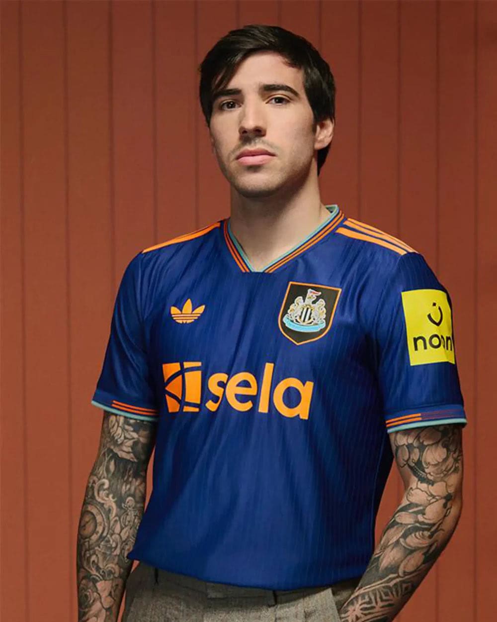 Sandrothd-RESIZE | AE JERSEY Portrait of a young man wearing a blue Newcastle United-style football jersey with orange trim, standing against an orange wooden backdrop.