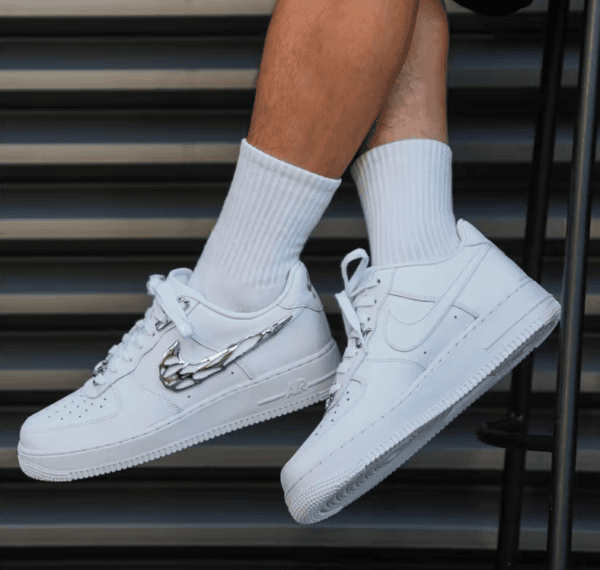 Pair of white Nike sneakers with a chrome wing design on the side, worn with white crew socks, resting on metal steps.