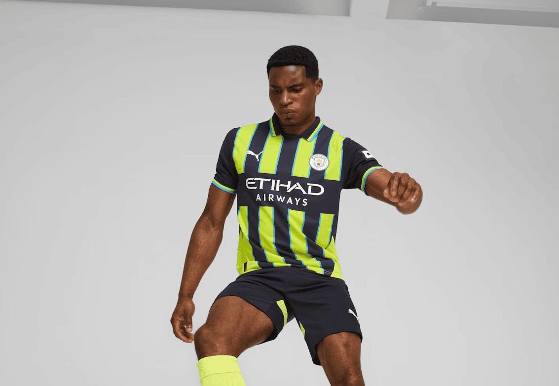 Screenshot_2024-09-04_181505 | AE JERSEY Soccer player in neon green and navy Man City away kit performing a kick on a light gray background, focused expression.