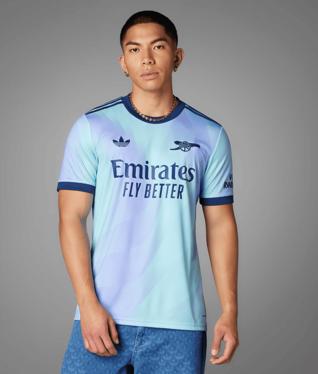 Screenshot_2024-09-18_194256_d6406abb-32a6-40d1-a824-5c66eea8548b | AE JERSEY Young man modeling a light-blue Arsenal away football jersey with 'Emirates Fly Better' on a gray studio background.