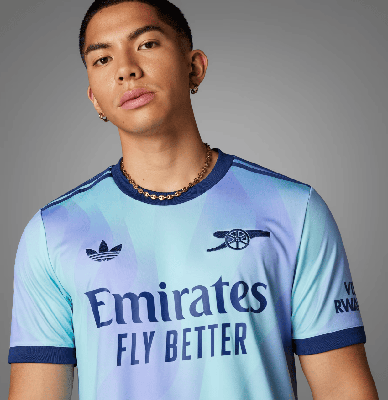 Screenshot_2024-09-18_194347_e9dc364f-38da-4e2e-be9b-9bf67388a332 | AE JERSEY Portrait of a young man wearing a light blue gradient football jersey with the words 'Emirates Fly Better', plus a gold chain and hoop earrings, against a gray background.