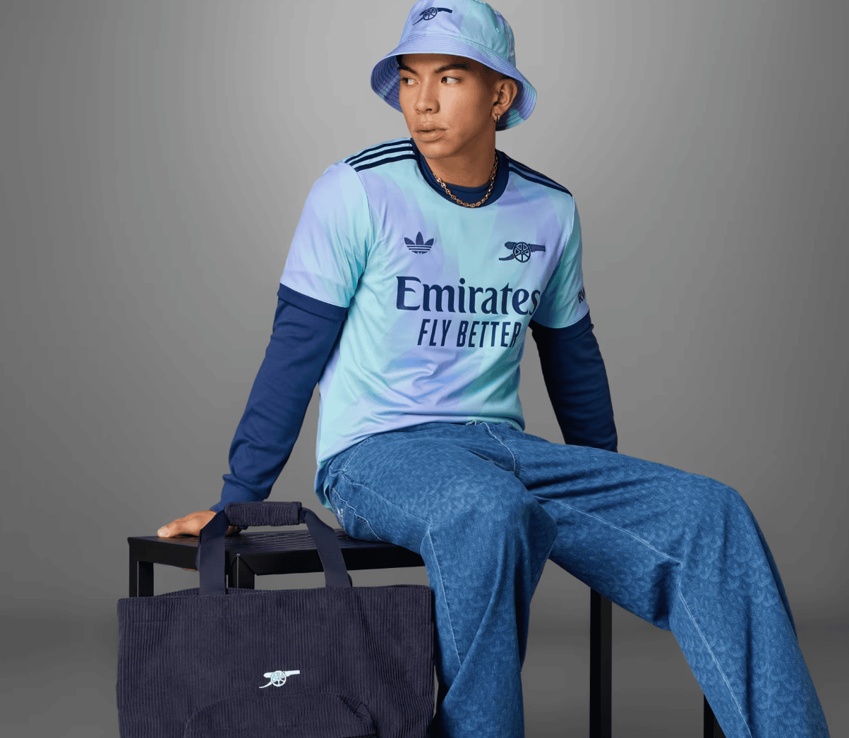 Screenshot_2024-09-18_194416_afaf008d-62d6-46a0-b159-846a3fd22383 | AE JERSEY Person in a pastel blue Arsenal jersey and bucket hat sits on a black stool with a navy tote bag beside them.