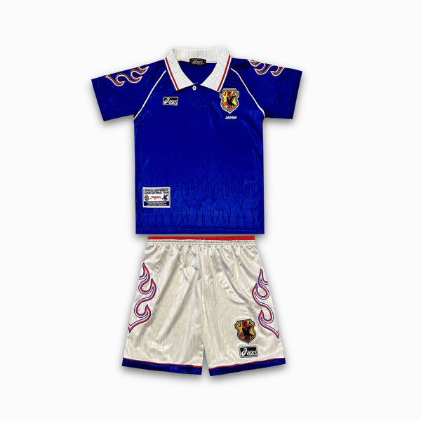 Blue soccer jersey with Japanese crest and white collar paired with white shorts featuring red-blue flame motifs and team badges, laid flat as a set for kids.