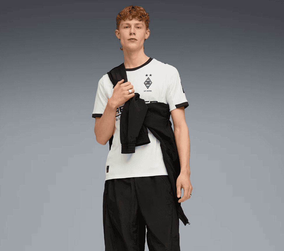 Screenshot_2025-07-16_205619 | AE JERSEY Male model wearing a white soccer jersey with crest, black shorts, and a black jacket draped over his shoulder against a gray gradient backdrop.