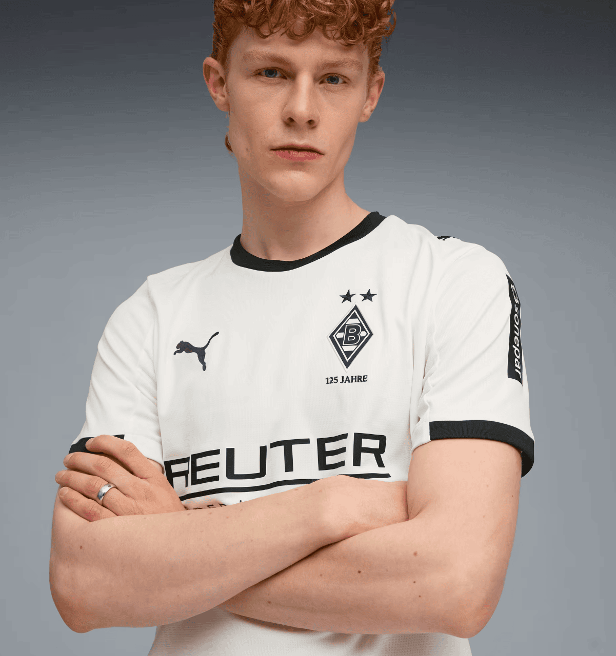 Screenshot_2025-07-16_205631 | AE JERSEY Portrait of a young man with red hair and a serious expression, arms crossed, wearing a white Borussia Mönchengladbach jersey against a gray gradient backdrop.