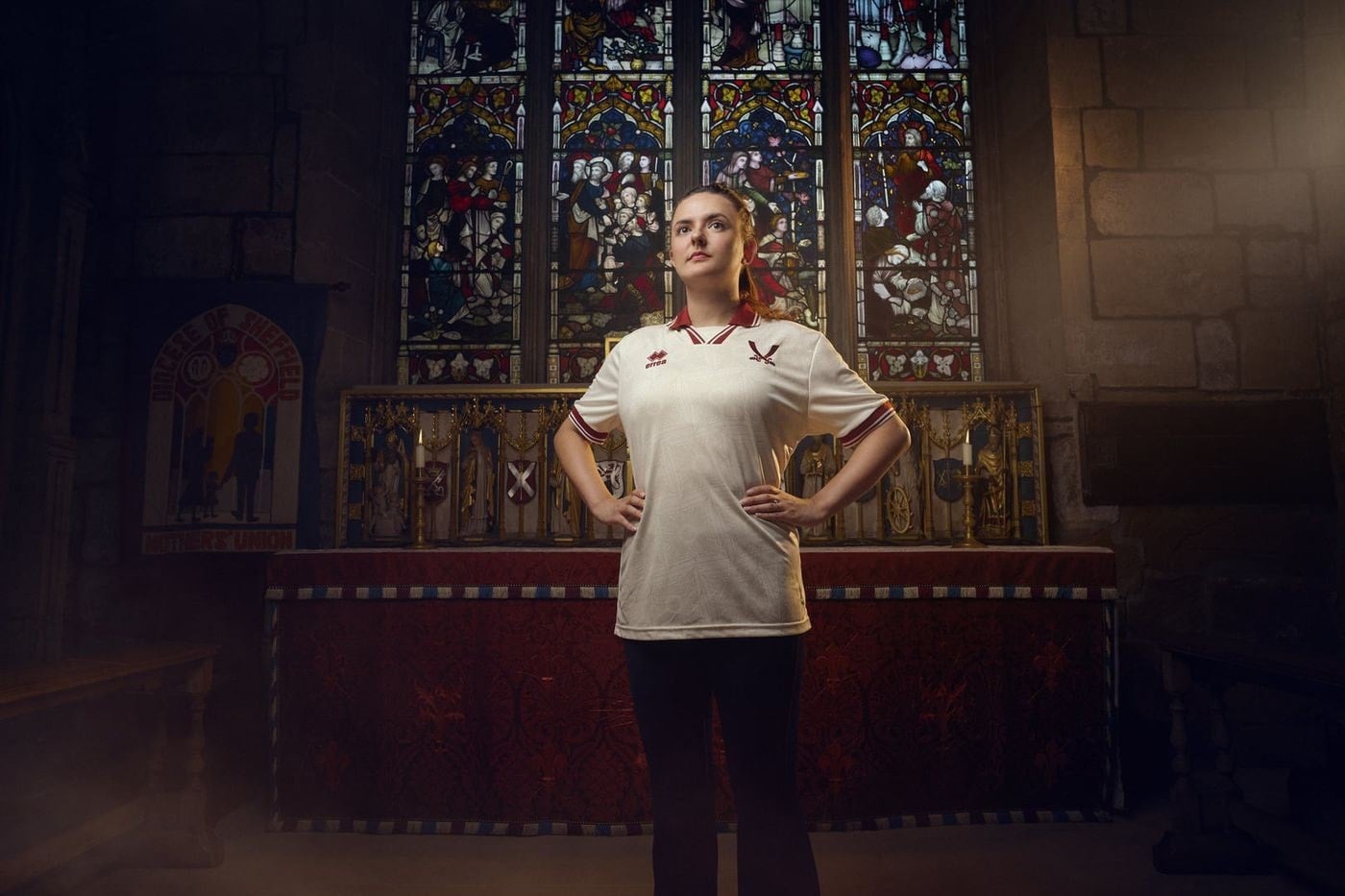 SjUYDN5mucLpZKW | AE JERSEY Woman in a white polo stands with hands on hips in a church, in front of an ornate altar and stained-glass window in the background.