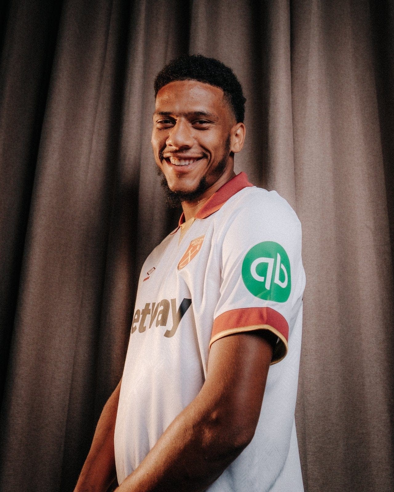 TPUw9lQA2bToUWU | AE JERSEY Smiling young man in a white team jersey with red trim, standing in front of dark stage curtains.