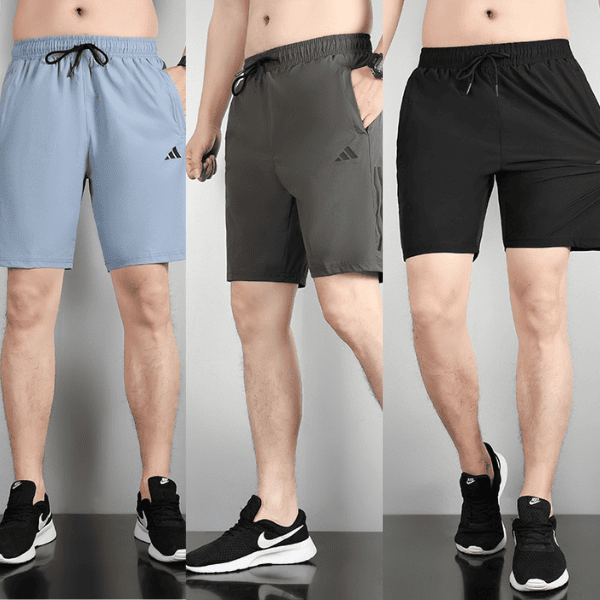 Three male models stand side by side wearing knee-length athletic shorts in light blue, gray, and black with drawstrings and small brand logos on the left thigh; black athletic shoes complete the look.