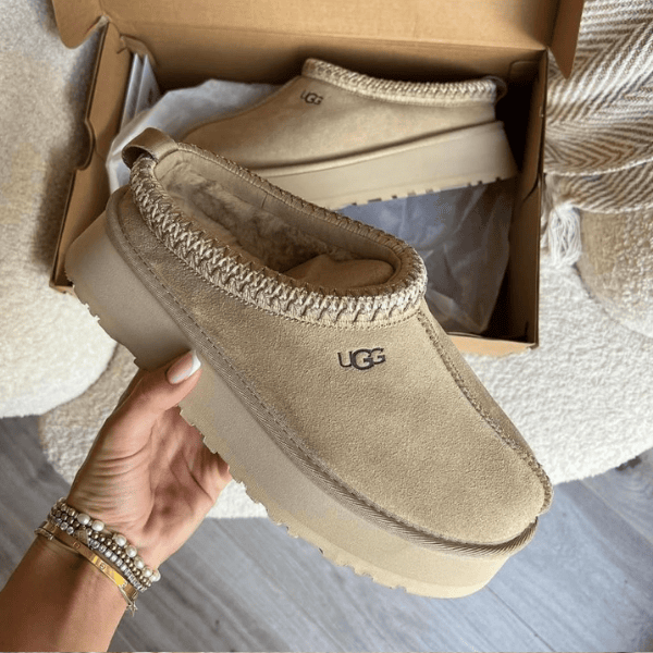 Beige UGG moccasin-style slipper with fuzzy lining held in hand, opened box in background