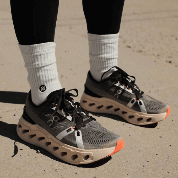 Pair of gray running shoes with a distinctive hollow-cut sole, black and orange laces, worn with white crew socks on pavement.