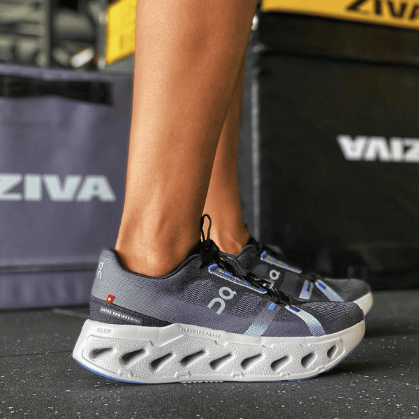 Person wearing dark gray On running shoes with white cloud-like sole standing on a gym floor.