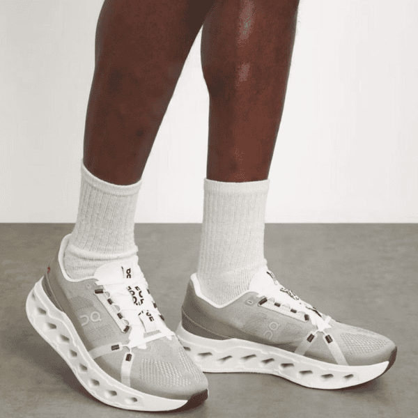Gray running shoes with white sole and sculpted cutouts, worn with white crew socks on a person’s feet, indoor setting.