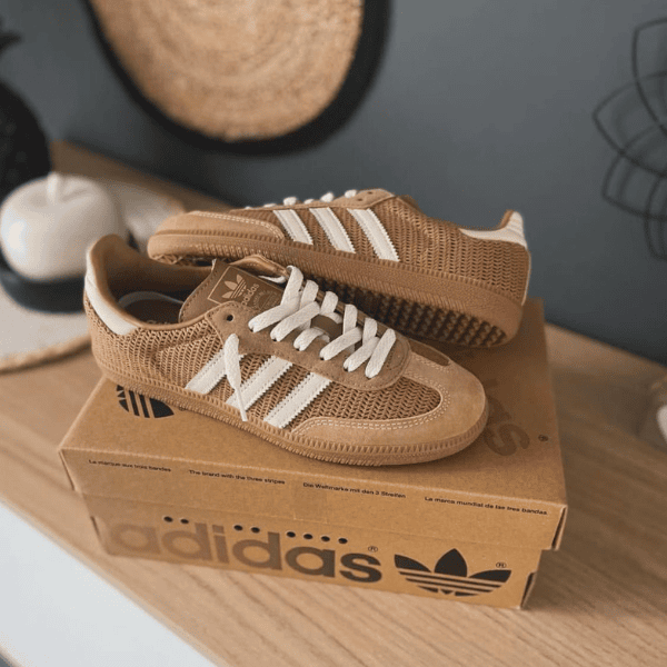 Brown Adidas sneakers with white stripes resting on a cardboard Adidas box on a wooden surface indoors.