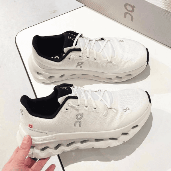 Pair of white running shoes with black inner lining displayed on a white surface next to a gray box, featuring the On logo on the sides and midsole holes for cushioning.